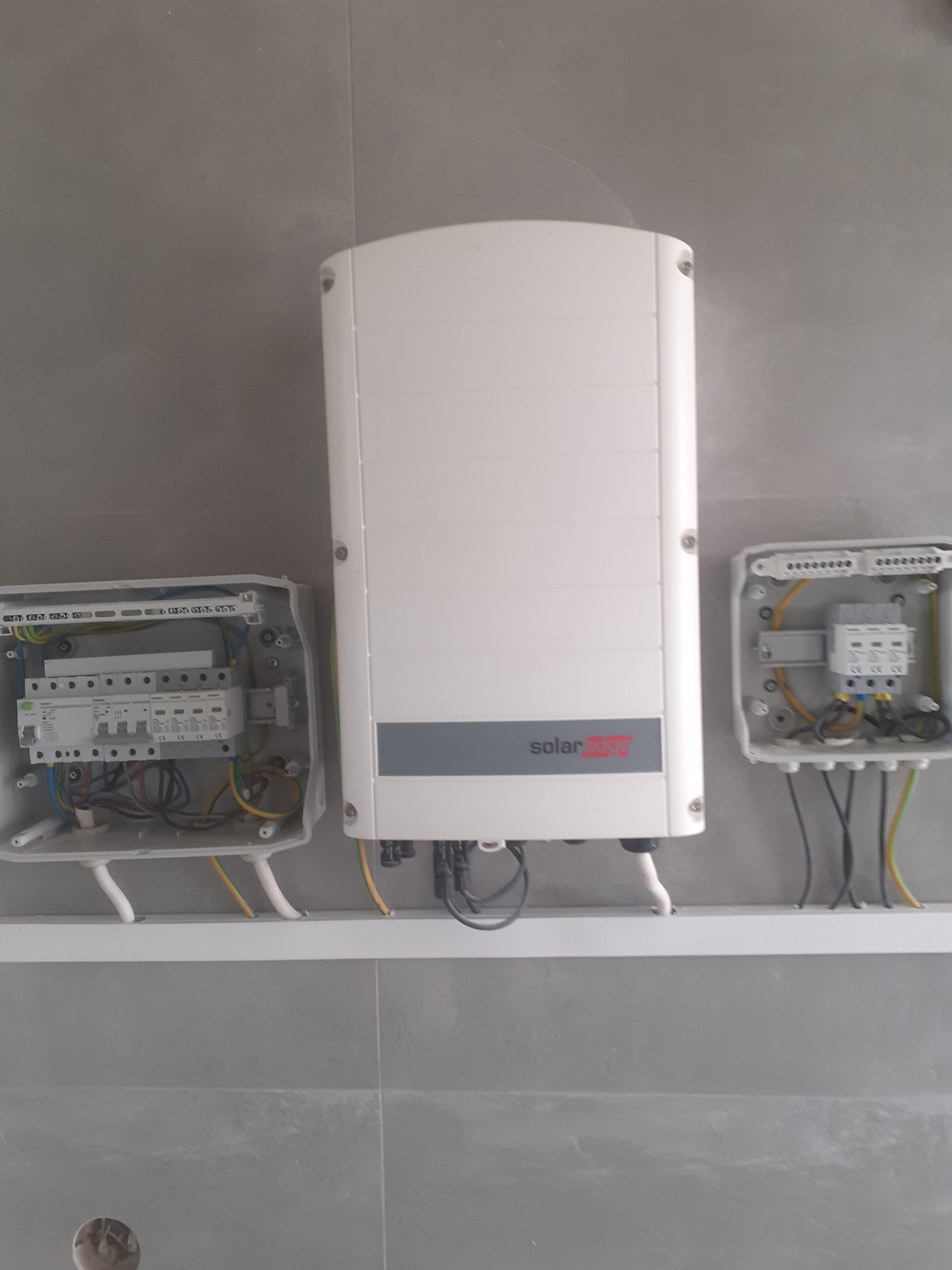 Zamontowany biały falownik SolarEdge z szarym paskiem, obok otwarte skrzynki elektryczne z widocznymi przewodami i bezpiecznikami, na szarej ścianie.