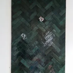 Ściana wyłożona błyszczącymi, ciemnozielonymi płytkami w układzie jodełki, z wystającymi kablami elektrycznymi w miejscu gniazdka.