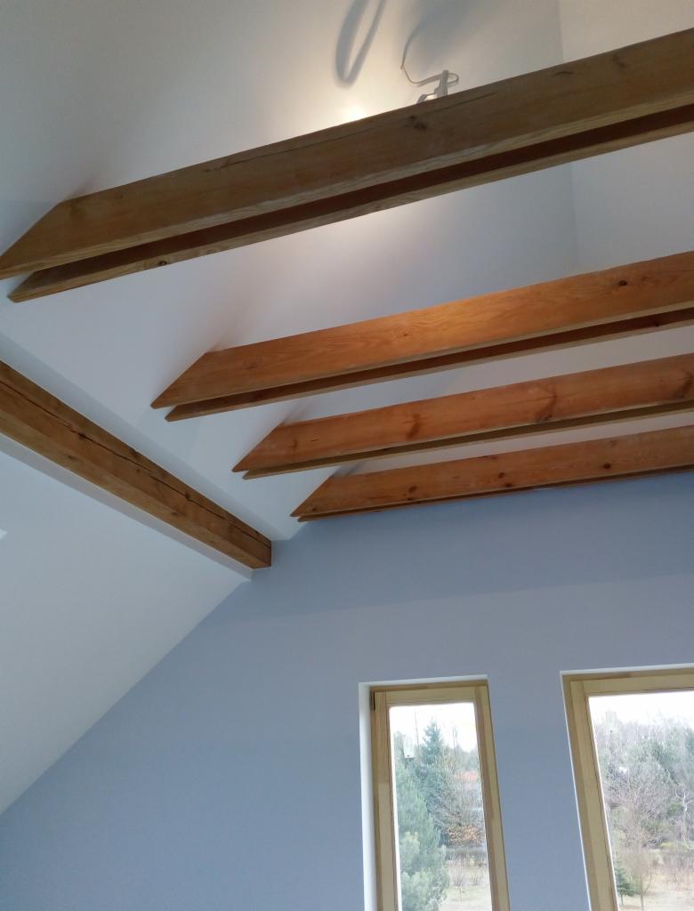 Jasne wnętrze z drewnianymi belkami stropowymi, szara ściana, dwa okna z widokiem na zewnątrz, oświetlenie sufitowe.