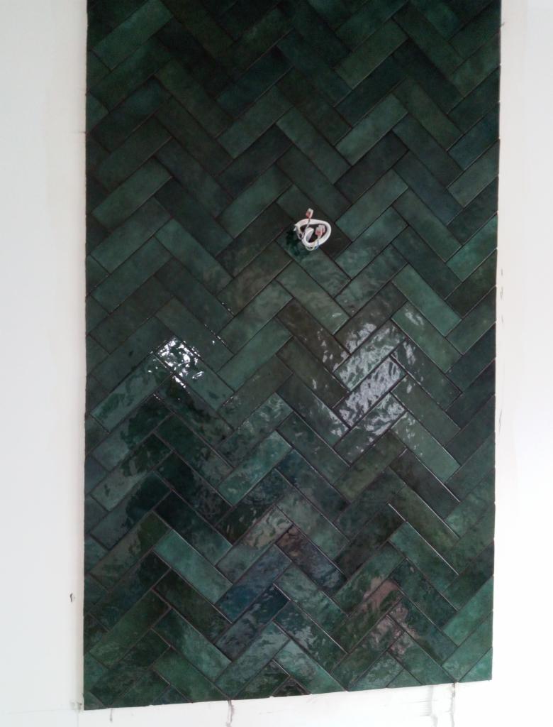 Ściana wyłożona błyszczącymi, ciemnozielonymi płytkami w układzie jodełki, z wystającymi kablami elektrycznymi w miejscu gniazdka.