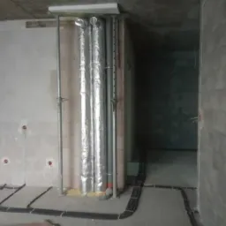Surowe wnętrze w trakcie prac wykończeniowych: widoczne kable elektryczne ułożone na podłodze, puszki instalacyjne w ścianie z betonu komórkowego oraz rury wentylacyjne w izolacji, podtrzymywane...