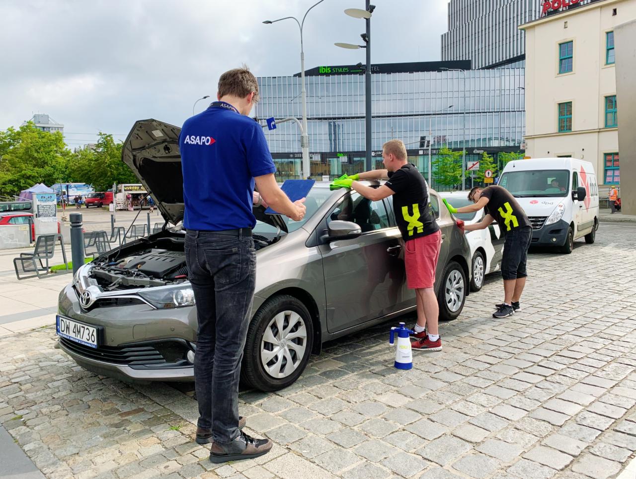 Zespół trzech osób w odzieży roboczej marki ASAPO czyści szary samochód Toyota na brukowanym placu miejskim, z otwartą maską i widocznym silnikiem, w tle budynek Ibis Styles i biały van DPD.