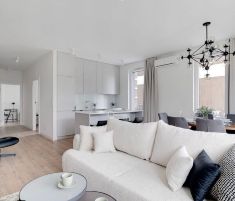 Jasne, nowoczesne wnętrze mieszkania z otwartą kuchnią, białym narożnikiem i designerską lampą wiszącą.