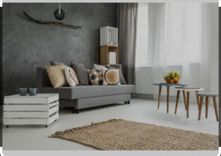 Szary salon z teksturowaną ścianą, szarą sofą z dekoracyjnymi poduszkami, białym stolikiem z drewnianych skrzynek, dywanem z juty i zestawem stolików kawowych.