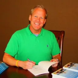 Uśmiechnięty mężczyzna w zielonej koszulce podpisuje egzemplarze swojej książki na drewnianym stole. W tle brązowa ściana.