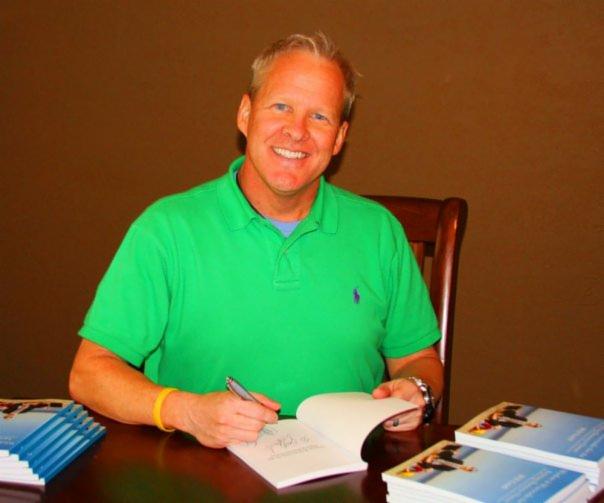 Uśmiechnięty mężczyzna w zielonej koszulce podpisuje egzemplarze swojej książki na drewnianym stole. W tle brązowa ściana.