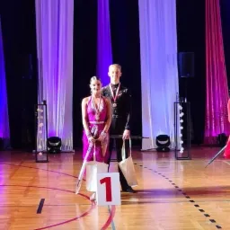 Para tancerzy w strojach galowych z numerem 1 na podeście, świętująca zwycięstwo na tle sceny z fioletowymi i białymi zasłonami.