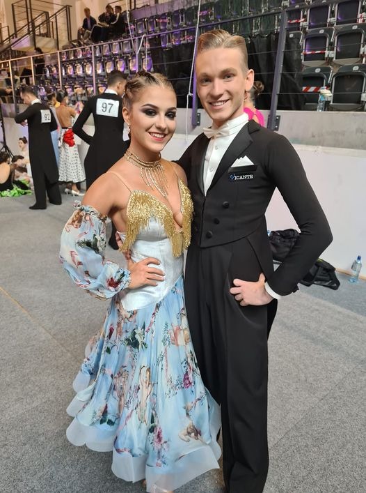 Para tancerzy w strojach galowych: kobieta w błękitnej sukni z kwiatowym wzorem i złotymi frędzlami, mężczyzna w czarnym fraku z białą muszką, uśmiechnięci, na tle widowni.