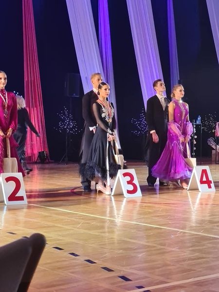 Tancerze w strojach konkursowych stojący na parkiecie z numerami 2, 3 i 4, oczekujący na ogłoszenie wyników zawodów.