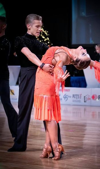 Para tancerzy w dynamicznej pozie na parkiecie: mężczyzna w czarnym stroju z kryształkami, kobieta w pomarańczowej sukience z frędzlami, wysokie obcasy, oświetlenie sceniczne, widoczny fragment...