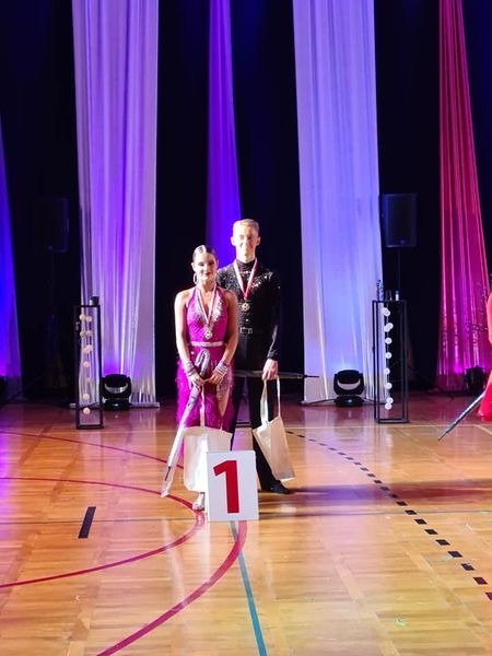 Para tancerzy w strojach galowych z numerem 1 na podeście, świętująca zwycięstwo na tle sceny z fioletowymi i białymi zasłonami.