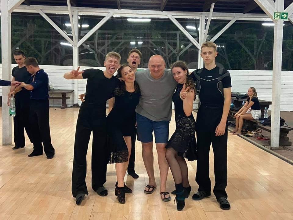 Grupa uśmiechniętych tancerzy pozuje do zdjęcia z instruktorem na parkiecie, w tle drewniana konstrukcja hali i inni uczniowie.