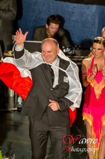 Mężczyzna w garniturze i pelerynie z flagą Polski macha ręką na parkiecie, obok kobieta w sukience tanecznej, w tle DJ.