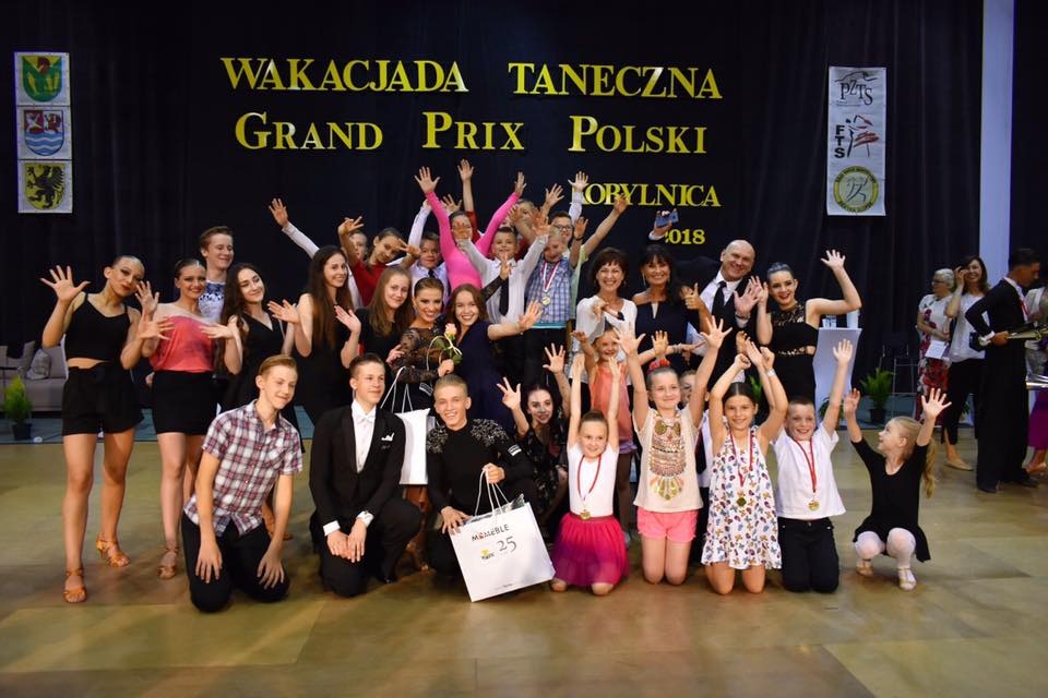 Grupa tancerzy w różnym wieku, w strojach tanecznych i eleganckich, pozuje z podniesionymi rękami na tle baneru 'Wakacjada Taneczna Grand Prix Polski', niektórzy z medalami, jeden trzyma torbę...