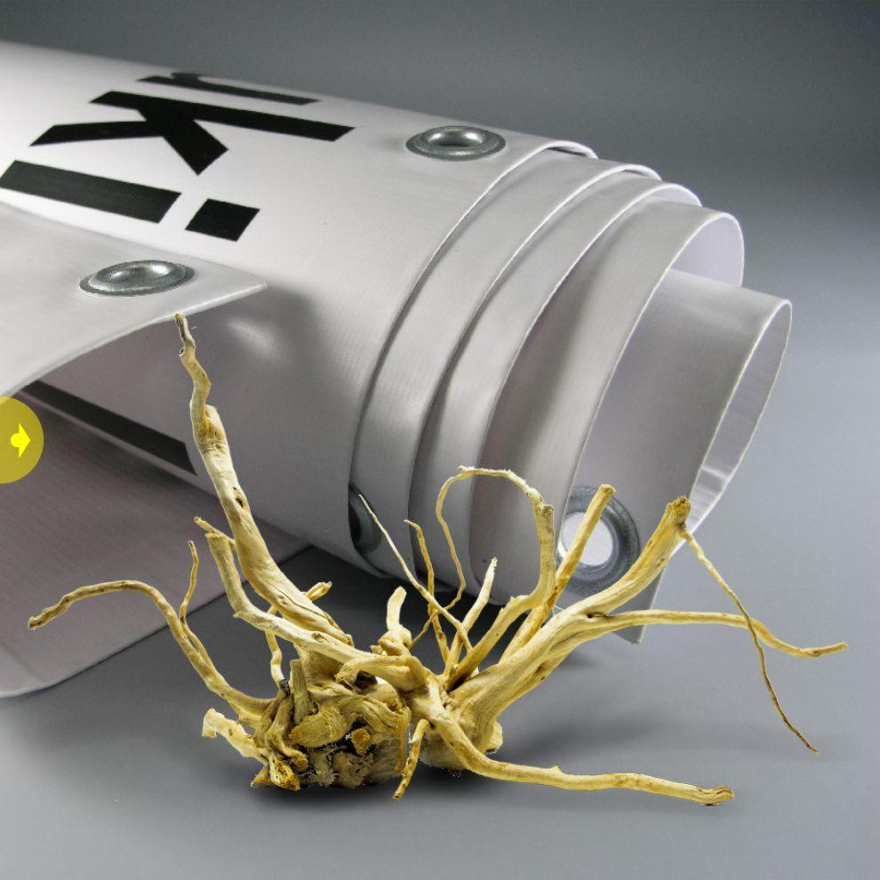 Zwinięty baner reklamowy z metalowymi oczkami, leżący obok dekoracyjnego korzenia na szarym tle.