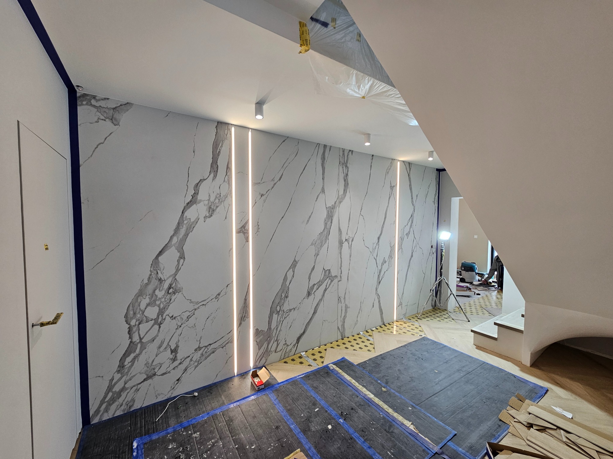 Ściana wykończona płytami imitującymi marmur z wkomponowanym oświetleniem LED, widoczne przygotowane podłoże pod parkiet i materiały budowlane na podłodze.