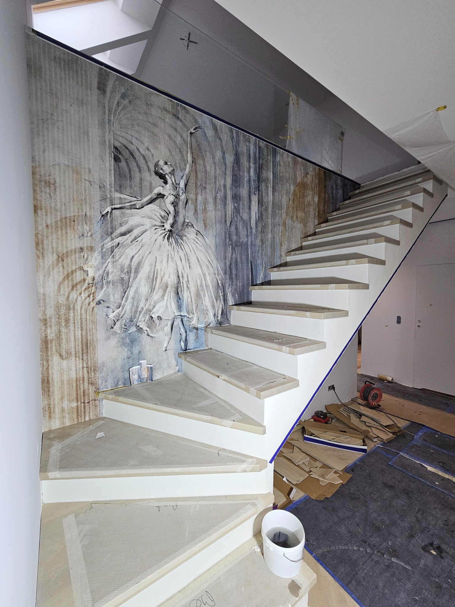 Montaż tapety z motywem baleriny na ścianie przy schodach, widoczne przygotowane stopnie schodów, narzędzia i materiały budowlane na podłodze.