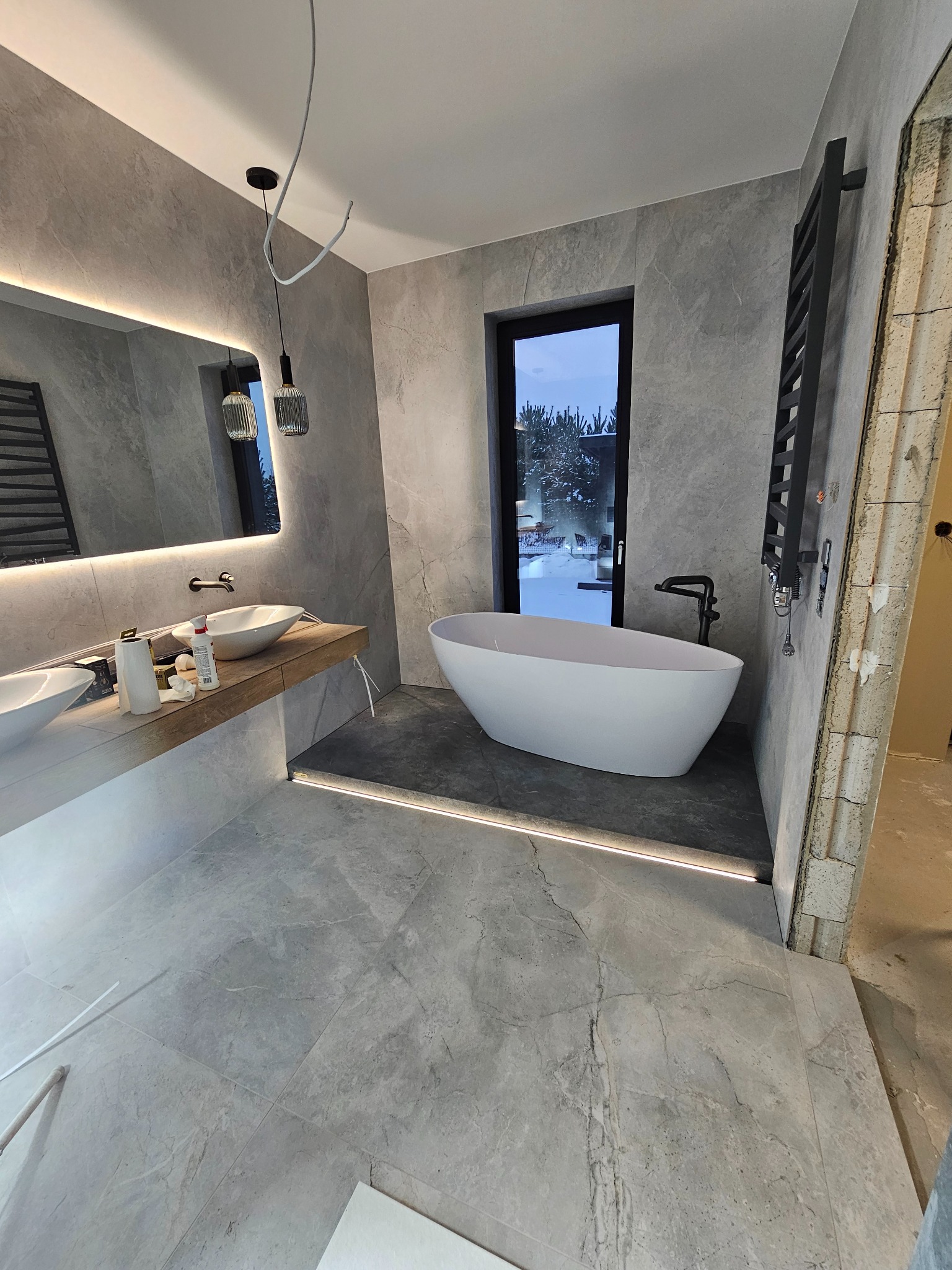 Nowoczesna łazienka z wolnostojącą wanną, szarą marmurową okładziną ścian i podłogi, podświetlanym lustrem i drewnianym blatem pod umywalki, widoczne niedokończone wykończenia ściany.