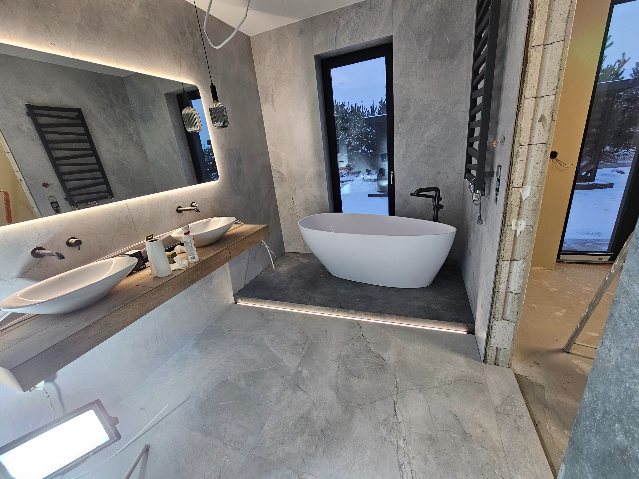Nowoczesna łazienka w trakcie wykańczania: szare płytki, wolnostojąca wanna, drewniany blat pod umywalkami, podświetlane lustro i widoczne prace budowlane przy drzwiach.