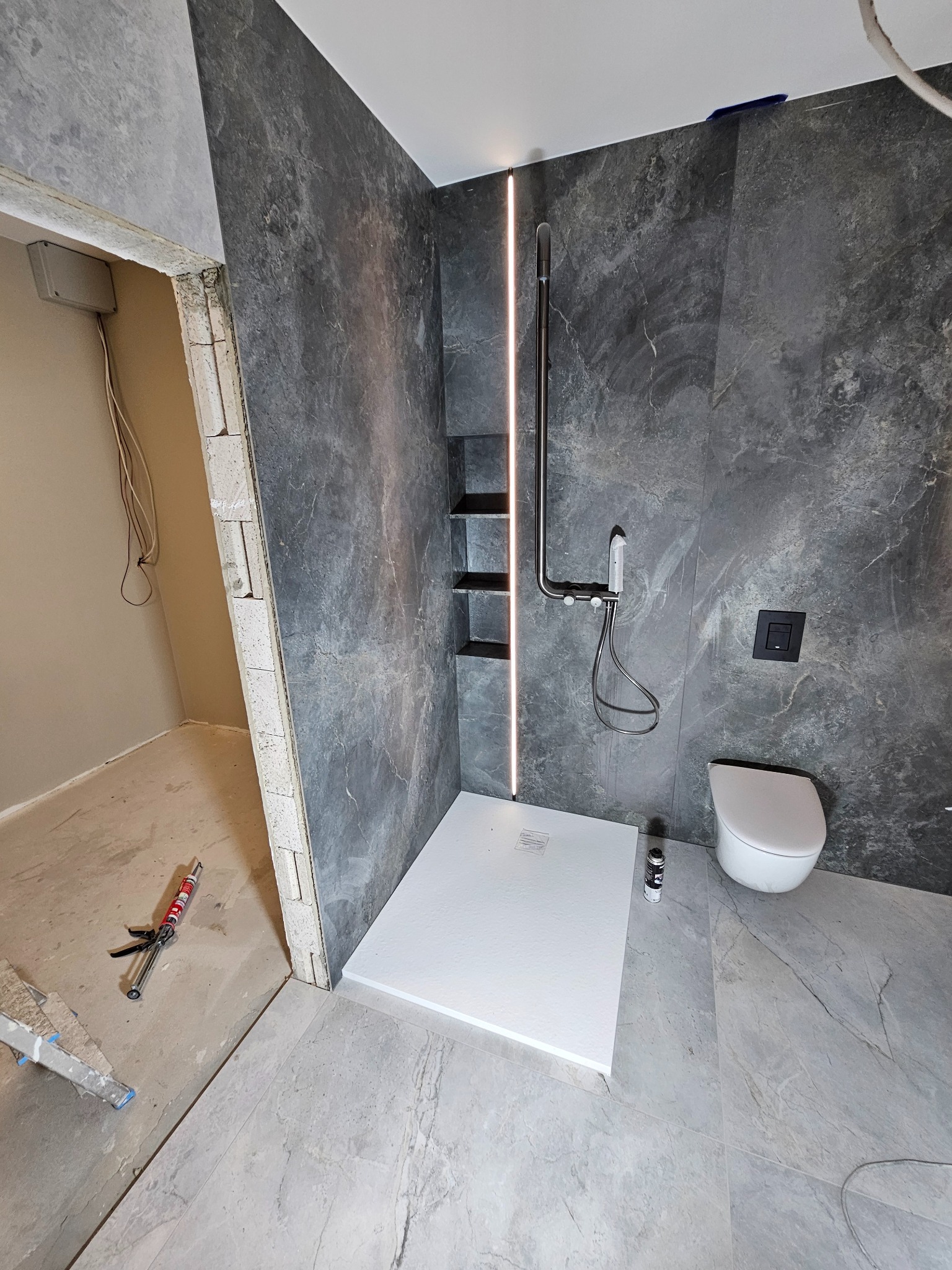 Remont łazienki w trakcie: marmurowe płytki ścienne z wnęką i oświetleniem LED, biały brodzik, wisząca toaleta, częściowo wykończone podłoże z narzędziami i silikonem w tle.