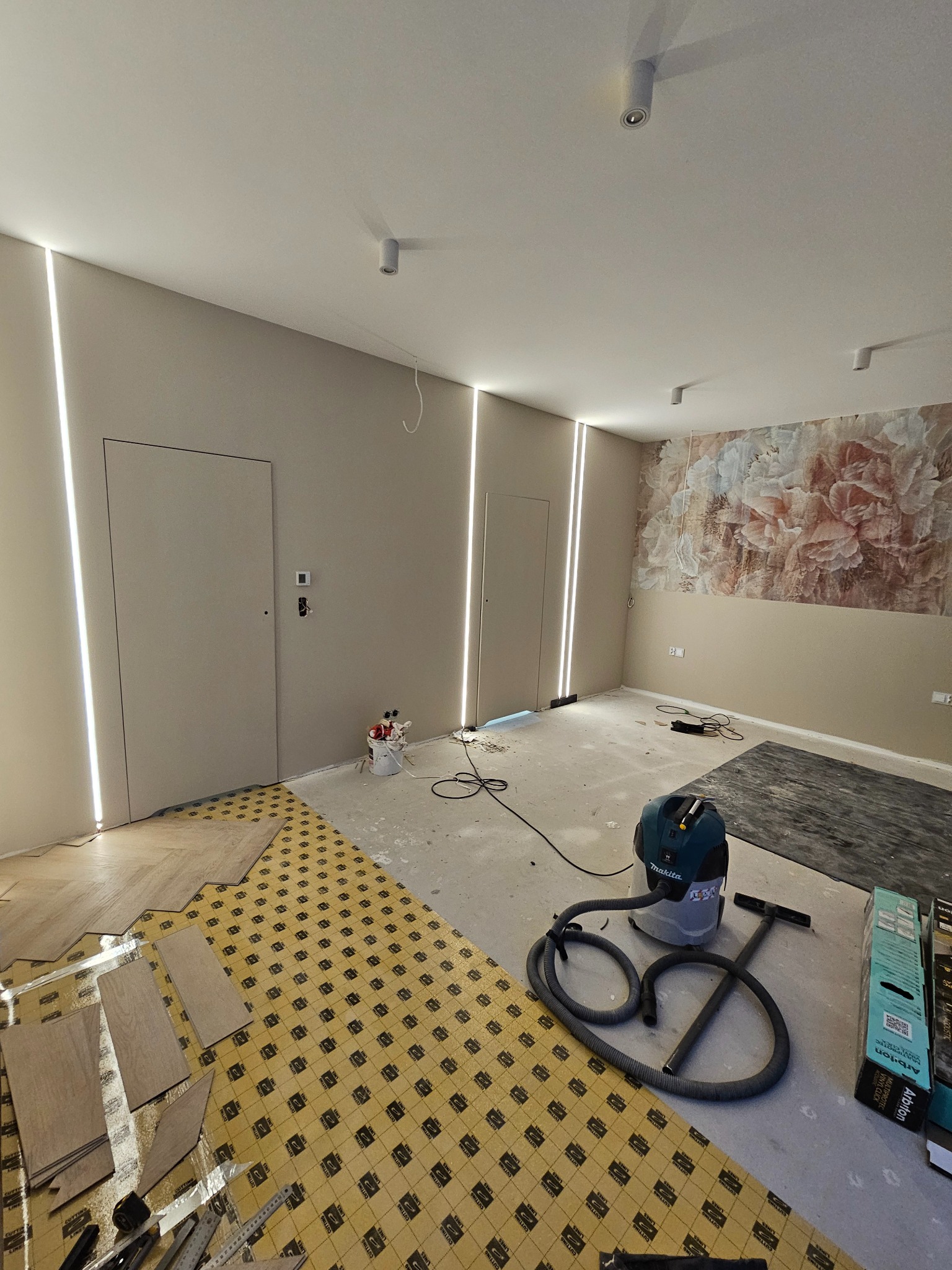 Wnętrze w trakcie układania podłogi z paneli, widoczny podkład, panele oraz odkurzacz przemysłowy Makita, ściany w kolorze beżowym z dekoracyjnym malowaniem kwiatowym i ukrytymi liniami świetlnymi.