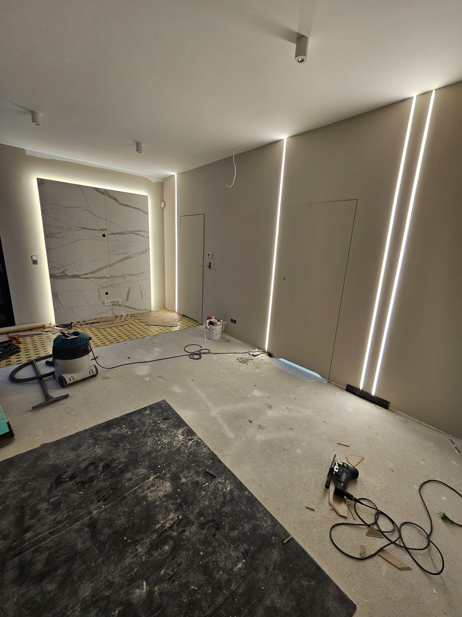 Wnętrze w trakcie remontu: widoczne pionowe pasy oświetlenia LED w ścianach, przygotowane podłoże pod parkiet, odkurzacz przemysłowy i szlifierka oscylacyjna. Fragment ściany wykończony płytami...