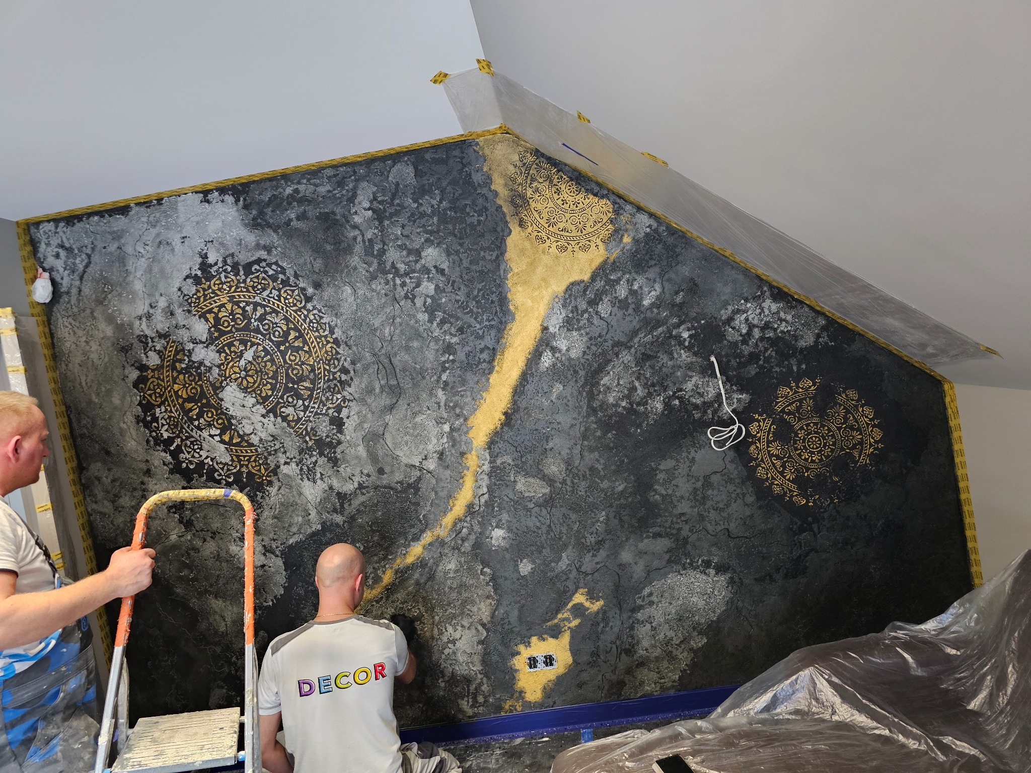 Dwóch mężczyzn na drabinach pracuje nad dekoracyjną ścianą z szaro-czarną teksturą, złotymi ornamentami i zabezpieczonymi taśmą narożnikami sufitu.
