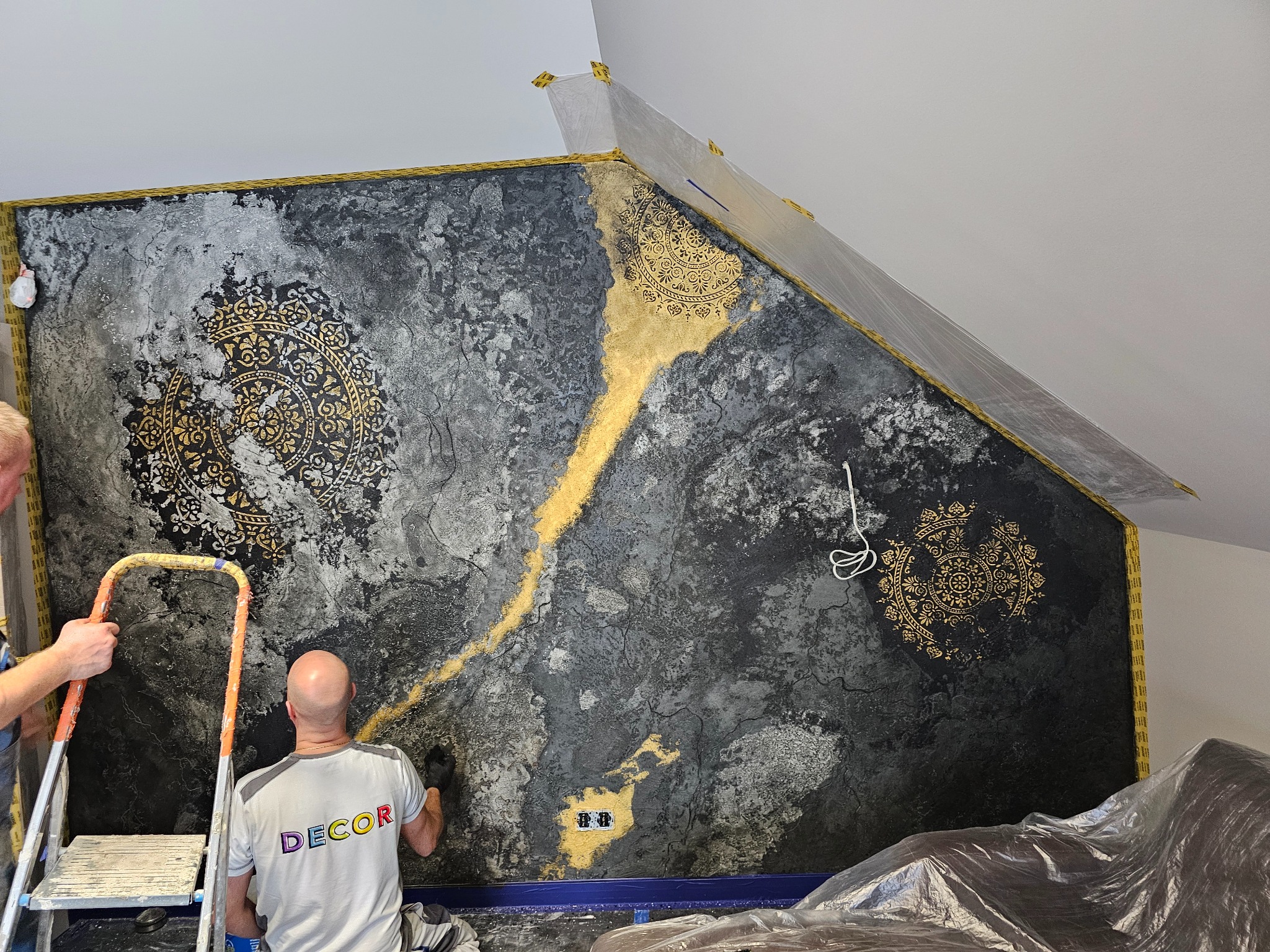Pracownik w koszulce z napisem DECOR nanosi gąbką dekoracyjną, czarno-złotą fakturę na ścianę w poddaszu, widoczna drabina i zabezpieczone taśmą krawędzie.