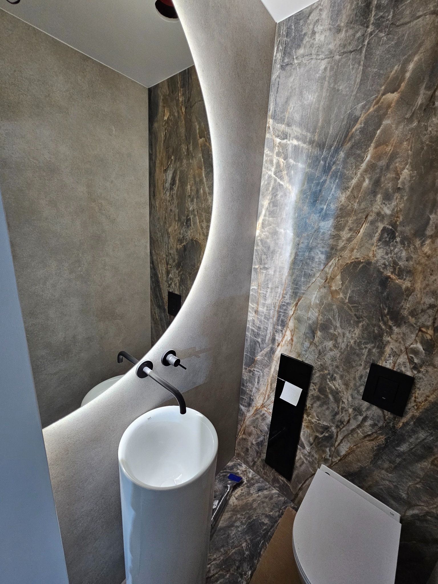 Nowoczesna łazienka z umywalką na postumencie, podświetlanym lustrem o nieregularnym kształcie i ścianami wykończonymi płytami imitującymi marmur w odcieniach szarości i brązu.