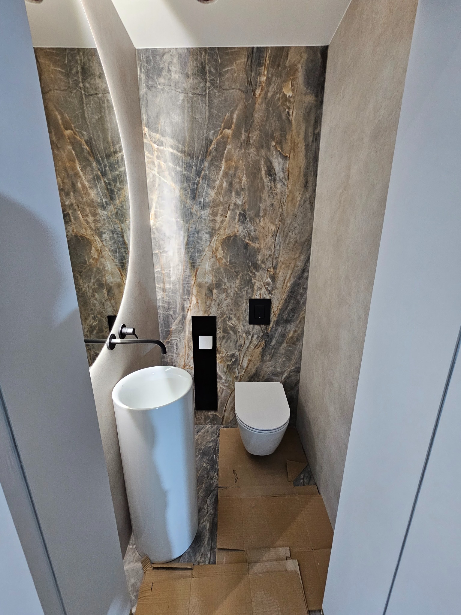 Nowoczesna łazienka z umywalką cylindryczną, lustrem o nieregularnym kształcie i marmurową ścianą akcentującą, widoczne zabezpieczenia podłogi kartonem.