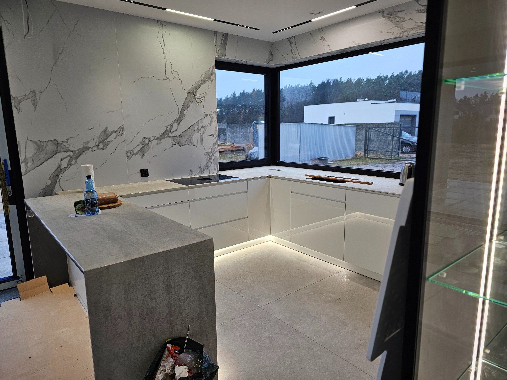 Nowoczesna kuchnia z białymi szafkami, marmurową ścianą i wyspą z blatem imitującym beton, widok na zewnątrz przez duże okno.