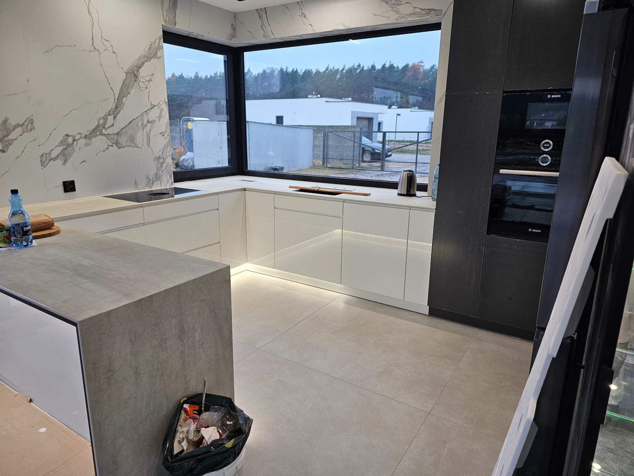 Nowoczesna kuchnia z białymi szafkami, szarą wyspą i marmurową ścianą, widok na posesję za oknem, ślady po remoncie widoczne na podłodze.