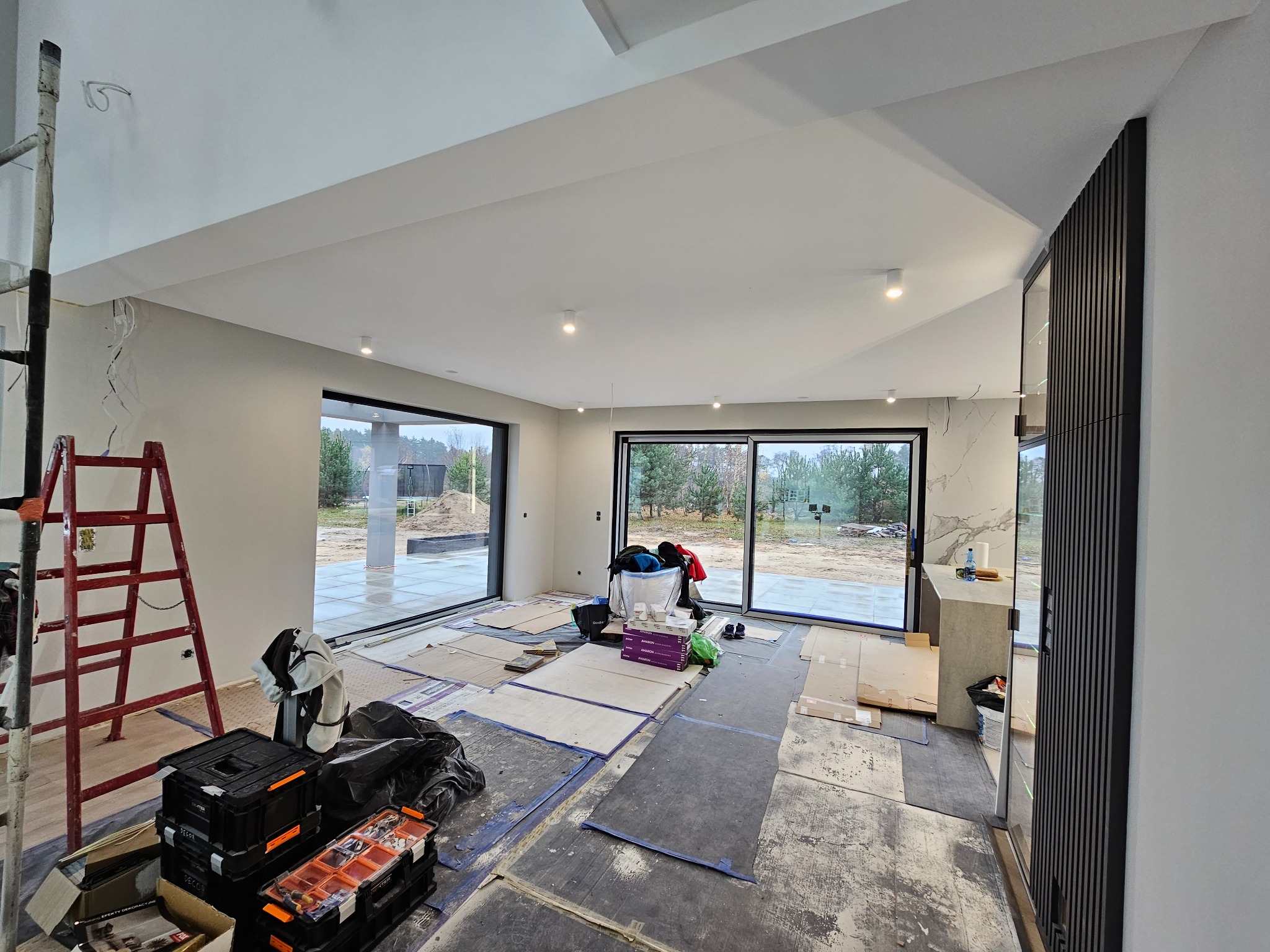 Wnętrze domu w Bydgoszczy w trakcie remontu, z widocznymi przygotowaniami do prac wykończeniowych: zabezpieczona podłoga, drabina malarska i skrzynki z narzędziami w rogu pomieszczenia, duże okna...