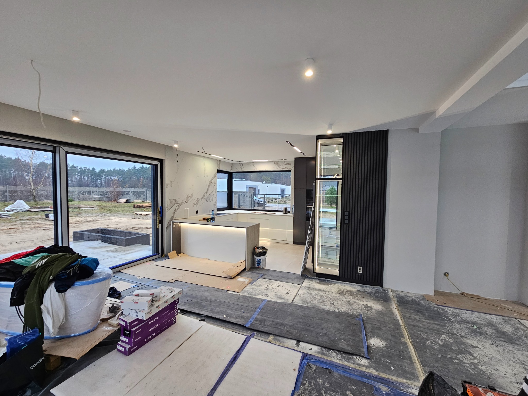 Wnętrze domu w trakcie remontu; widoczne przygotowane podłoże, płyty zabezpieczające, kartony z materiałami, duża przeszklona ściana z widokiem na zewnątrz.