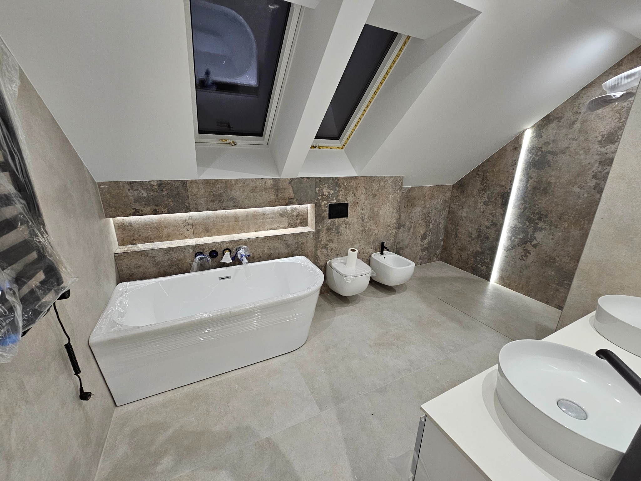 Wykończona łazienka na poddaszu ze skośnym sufitem i dwoma oknami dachowymi, z wanną w folii ochronnej, umywalką nablatową i bidetem. Ściany wyłożone płytkami imitującymi kamień, podświetlenie LED...