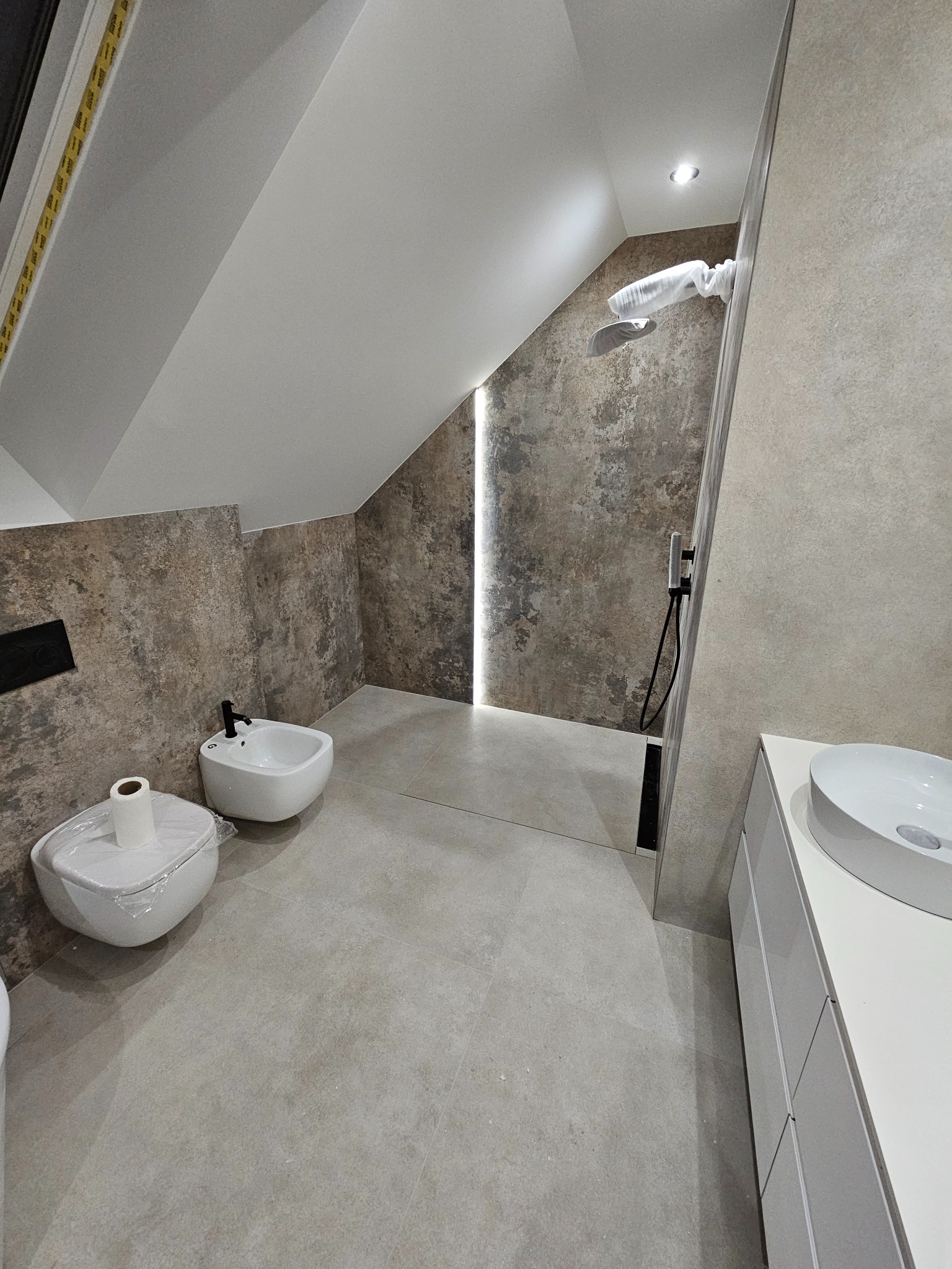 Nowoczesna łazienka na poddaszu z beżowymi płytkami imitującymi kamień, białą armaturą częściowo zabezpieczoną folią ochronną i oświetleniem LED w pionowym pasie.