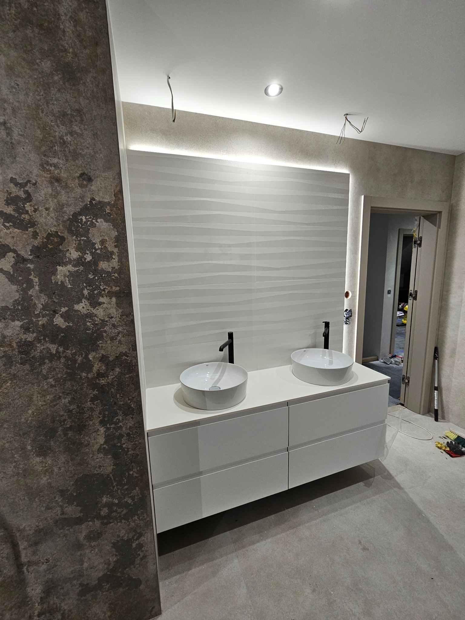 Nowoczesna łazienka w trakcie wykończenia: biała szafka z dwiema umywalkami nablatowymi i czarną armaturą, podświetlana ściana z dekoracyjnymi płytkami, widoczne przewody elektryczne i narzędzia...