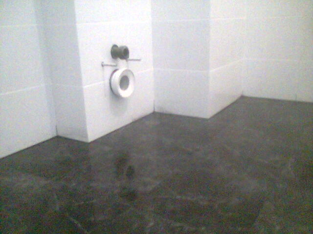 Świeżo położona, ciemna, błyszcząca posadzka w łazience z białymi, błyszczącymi płytkami na ścianach i wystającym przyłączem kanalizacyjnym.