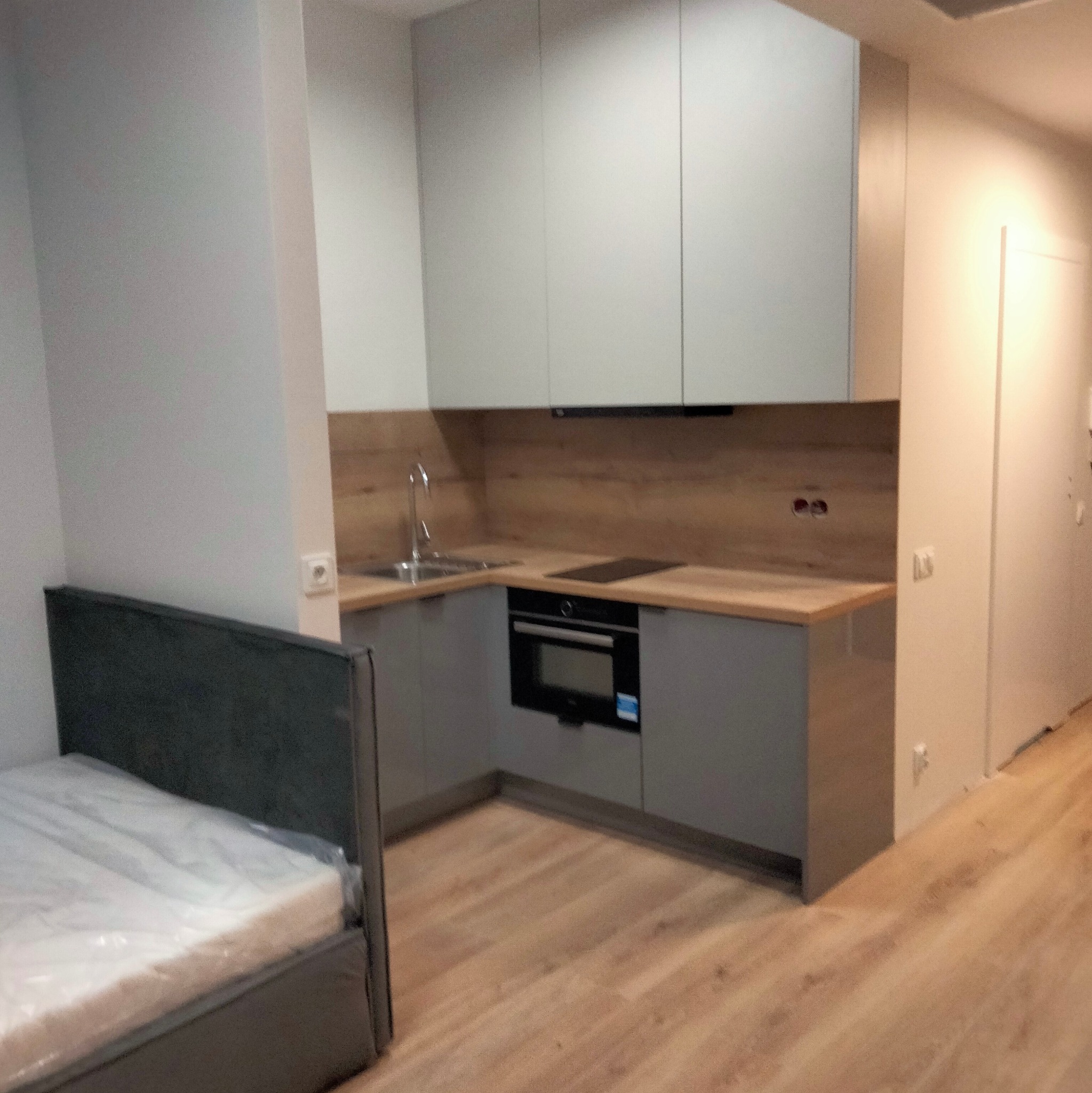 Mała kuchnia w aneksie: szare szafki na wymiar z drewnianym blatem i okładziną, obok łóżko z szarym zagłówkiem, podłoga drewnopodobna.