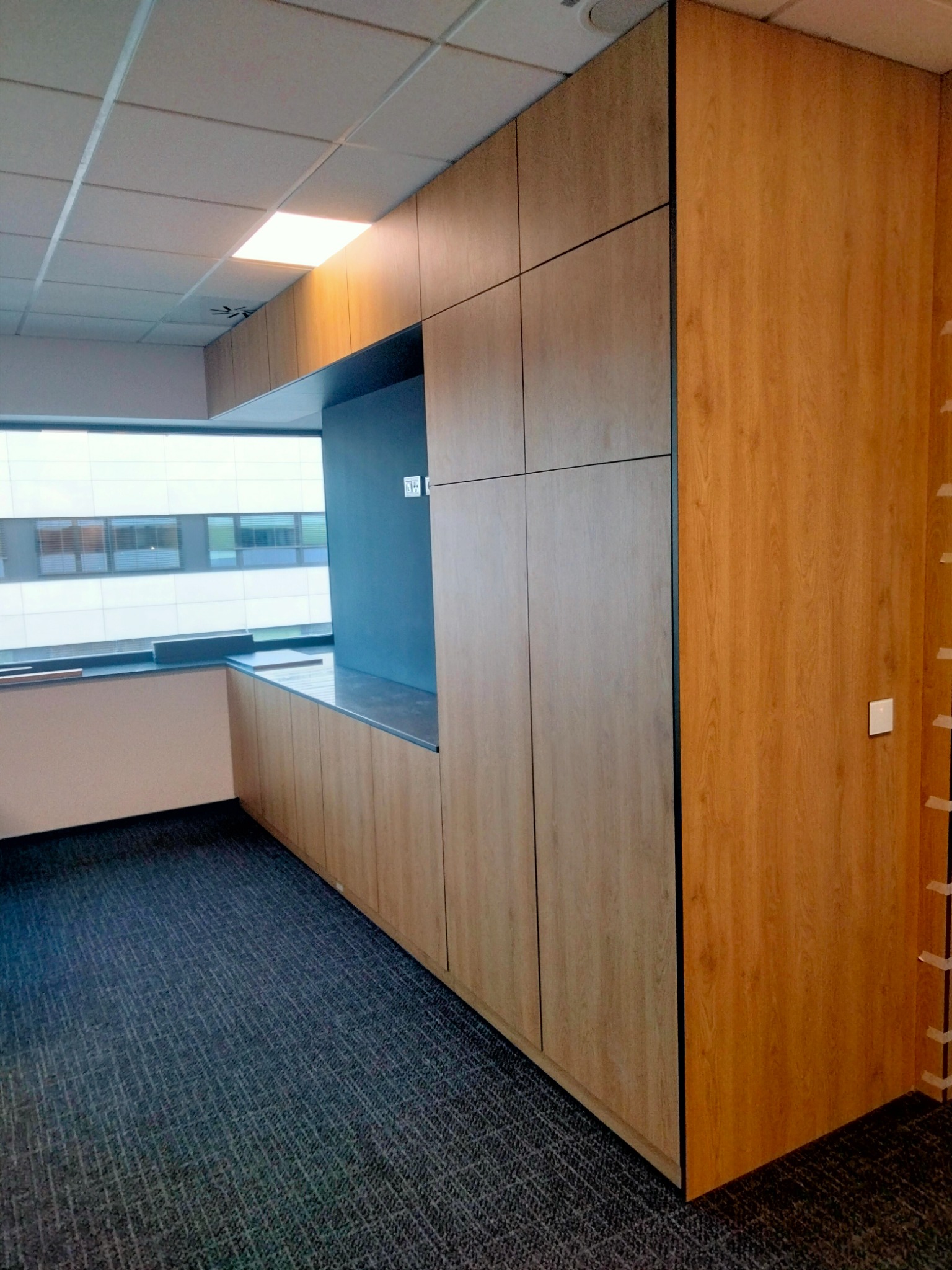 Drewniana zabudowa meblowa z szafkami i blatem w biurze, widok na okno z zewnątrz budynku.