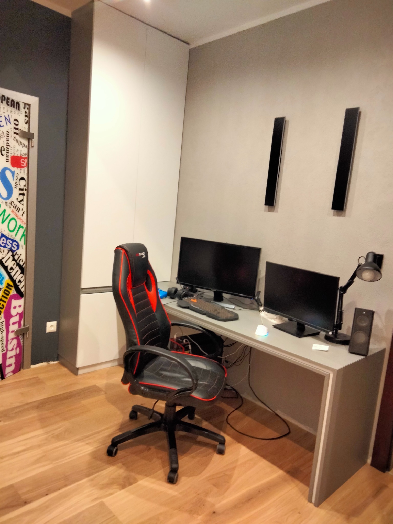 Pokój gracza z biurkiem, dwoma monitorami, krzesłem gamingowym z czerwonymi akcentami i szafą w zabudowie z białymi frontami, uzupełniony o drewnianą podłogę i dekoracyjną naklejkę na drzwiach.