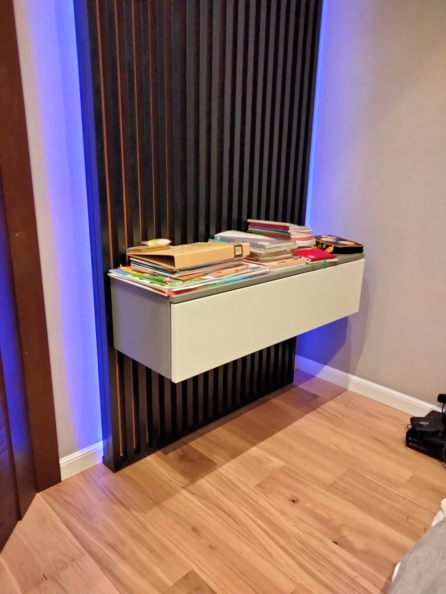 Biała, wisząca szafka z szufladą zamontowana na ścianie z drewnianymi lamelami podświetlonymi niebieskim światłem LED, na blacie sterta książek i zeszytów.