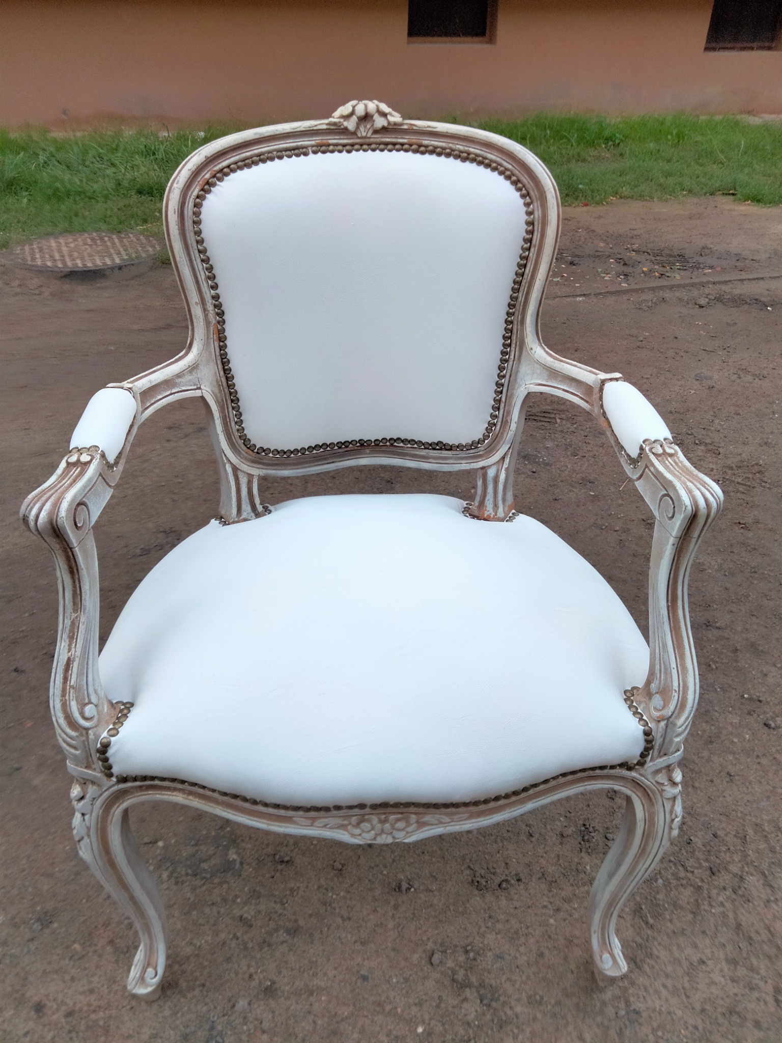 Biały, odnowiony fotel w stylu Ludwika XVI z dekoracyjnymi gwoździami, stojący na zewnątrz na ziemi.