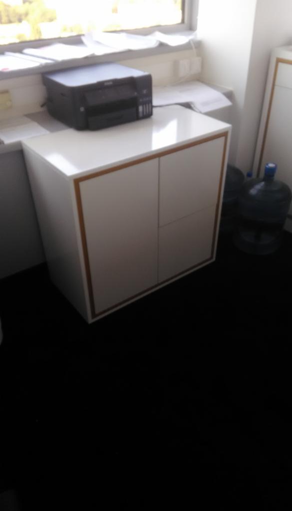 Biała szafka z dwoma drzwiczkami, obramowana cienką, drewnianą listwą, stojąca przy ścianie obok biurka z drukarką i butlami z wodą.