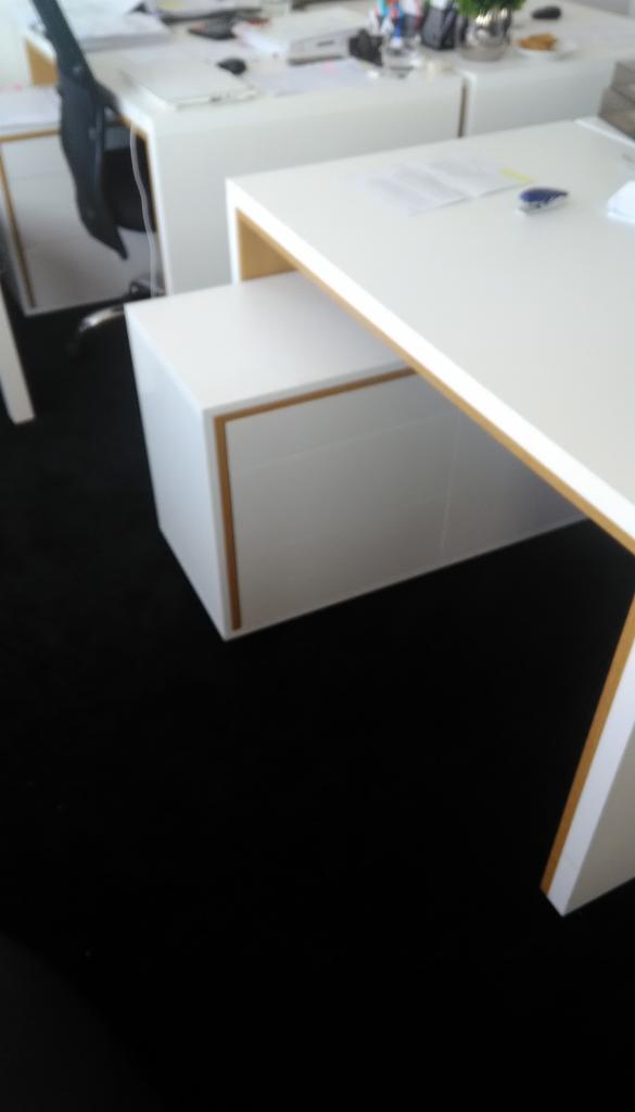 Białe biurko z szufladami w stylu minimalistycznym, z drewnianymi akcentami na krawędziach, stojące na czarnej wykładzinie.