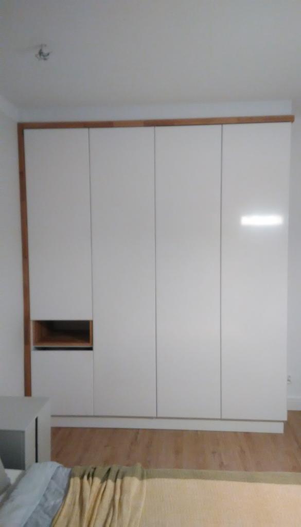 Wysoka, biała szafa z drewnianym obramowaniem i otwartą półką w dolnej części, widoczna w sypialni z fragmentem łóżka i stolika nocnego.