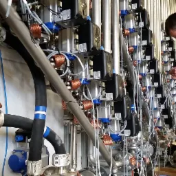 Pracownik w niebieskich spodniach roboczych przy ścianie z licznymi instalacjami i przewodami w obiekcie przemysłowym w Ostrowie Wielkopolskim, zajęty podłączaniem przewodów do automatyki...