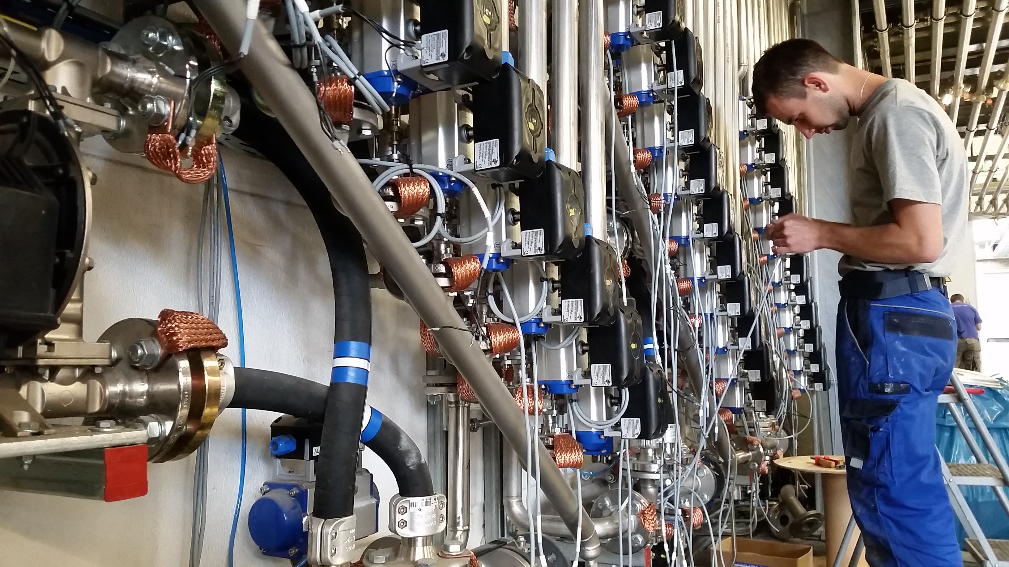 Pracownik w niebieskich spodniach roboczych przy ścianie z licznymi instalacjami i przewodami w obiekcie przemysłowym w Ostrowie Wielkopolskim, zajęty podłączaniem przewodów do automatyki...