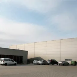 Nowoczesny budynek hali produkcyjnej z biurem, szara elewacja, brama wjazdowa, zaparkowane samochody, kostka brukowa na placu, pochmurne niebo.