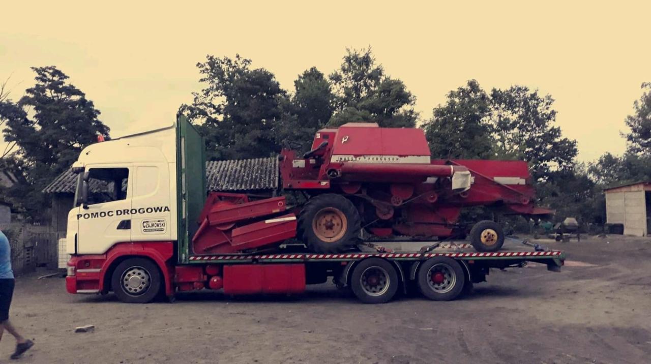 Ciężarówka pomocy drogowej transportuje czerwony kombajn rolniczy na platformie. Widoczne drzewa i budynki w tle, co sugeruje wiejski teren.